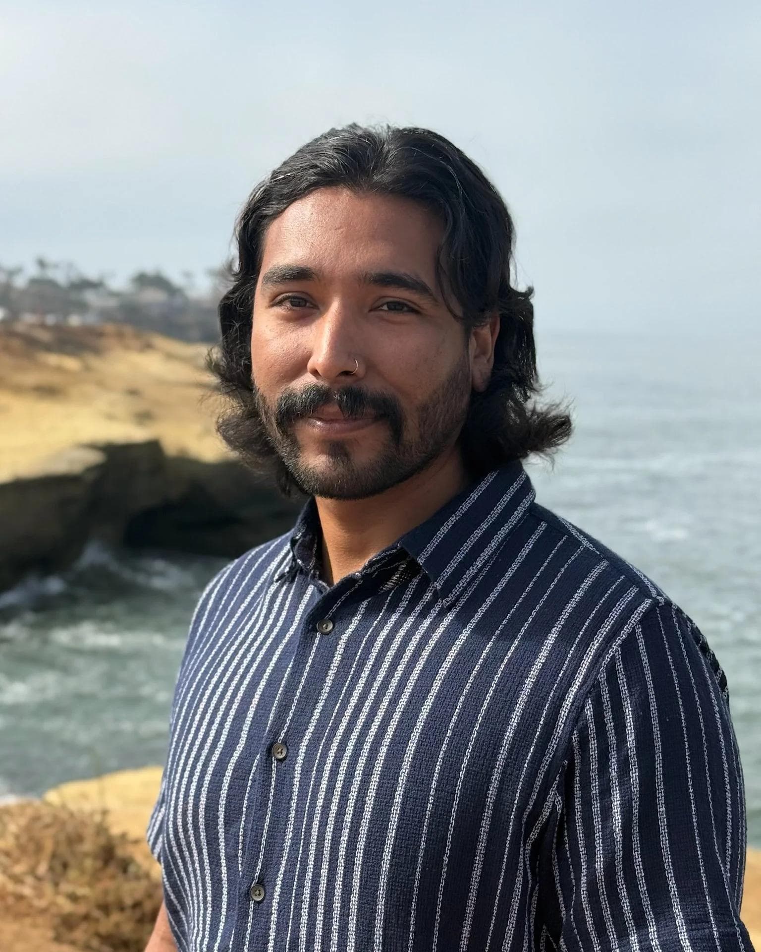 A headshot of Raul, a man with wavy dark hair, a beard, and a nose ring, wearing a blue and white striped shirt with a blurred coastal background.