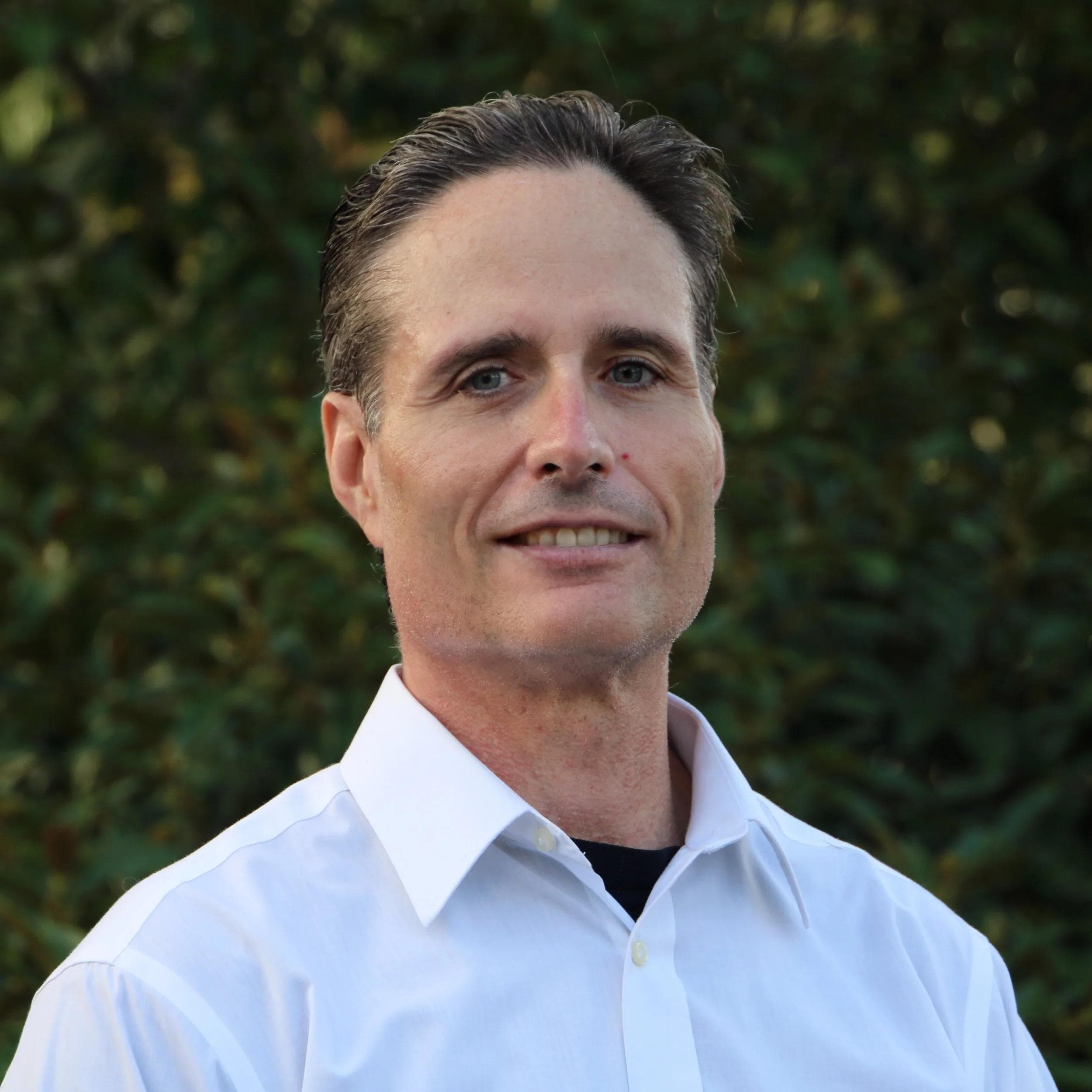 A portrait of Ed, a middle-aged man with short brown hair and a slight smile, wearing a white collared shirt against a blurred green foliage background.