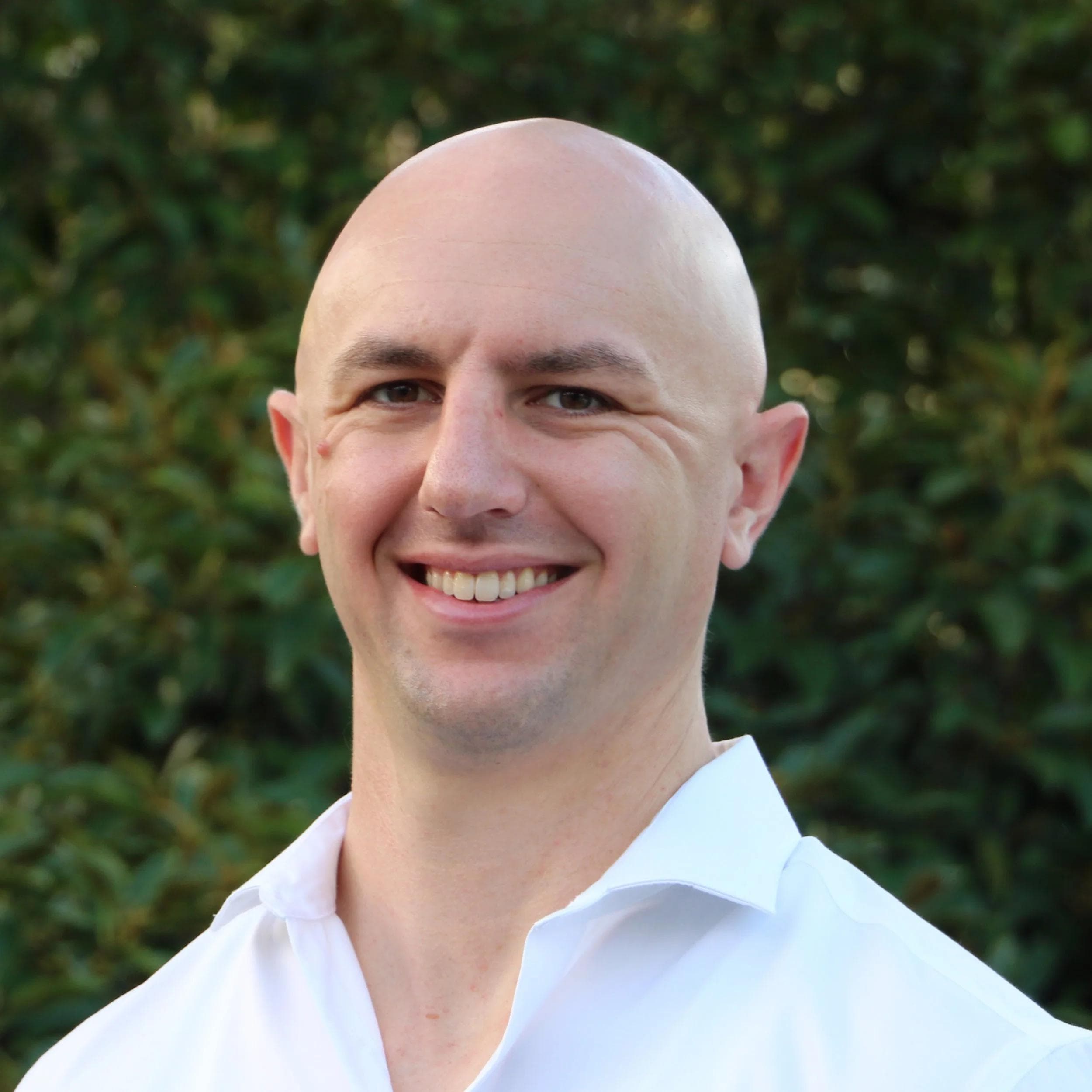Matt, a bald man with fair skin, smiles in a white shirt against a green leafy background.
