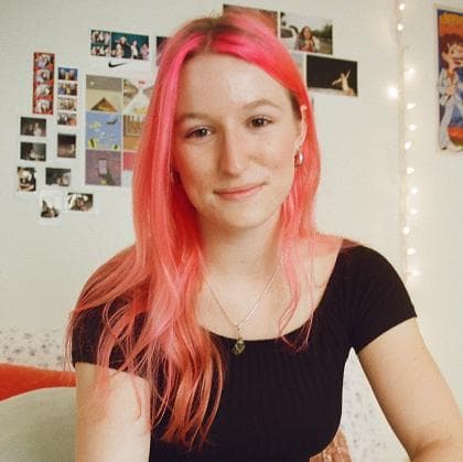 Grace, a young woman with bright pink hair and fair skin, smiles in a room decorated with photos and fairy lights.