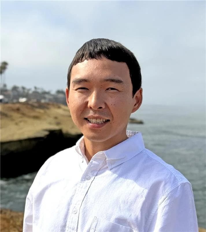 A headshot of Sandzhy, a man with short dark hair and a friendly smile, wearing a white button-down shirt while standing on a cliff in front of the ocean.