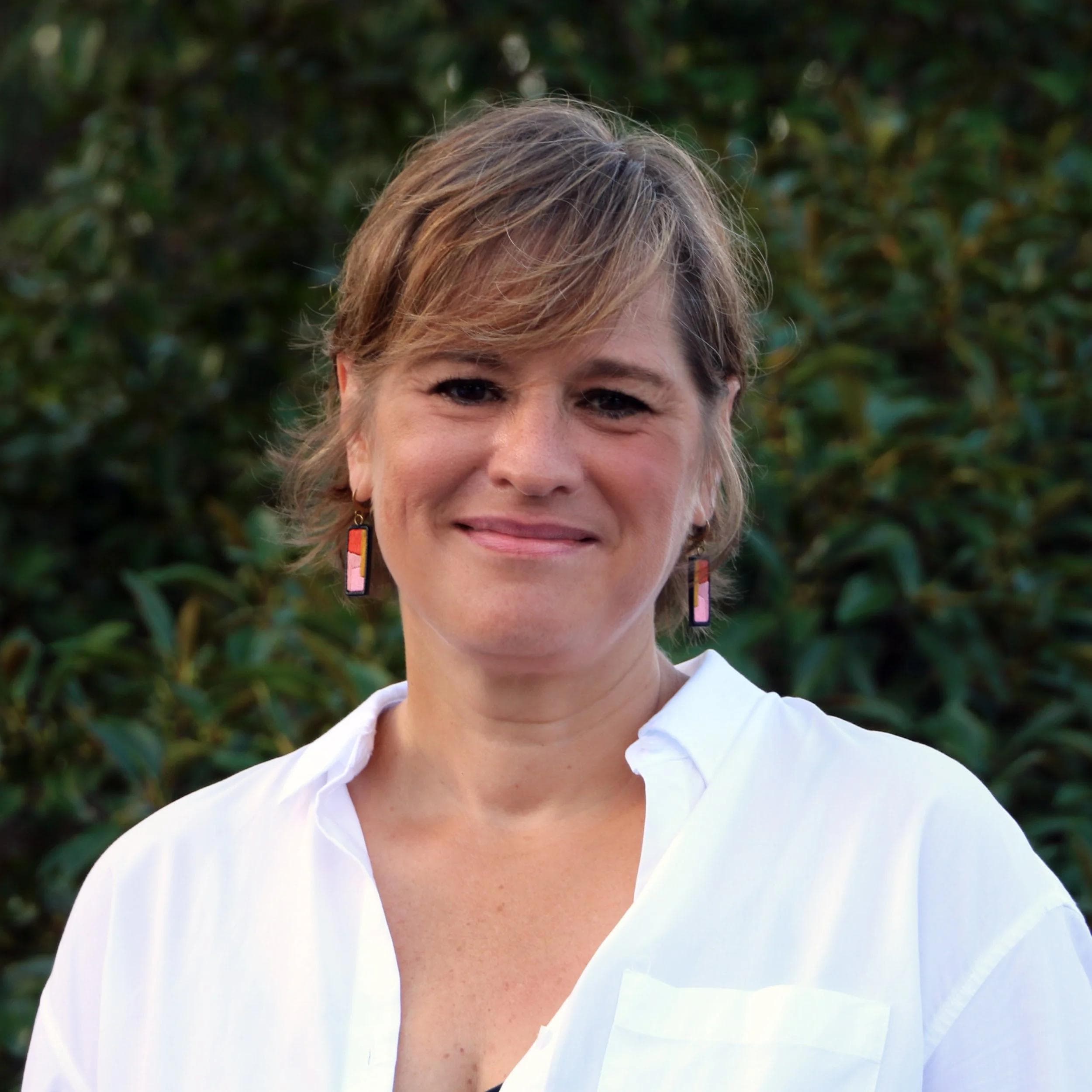 Heather, a woman with short brown hair and colorful earrings, smiles while wearing a white shirt against a leafy green background.